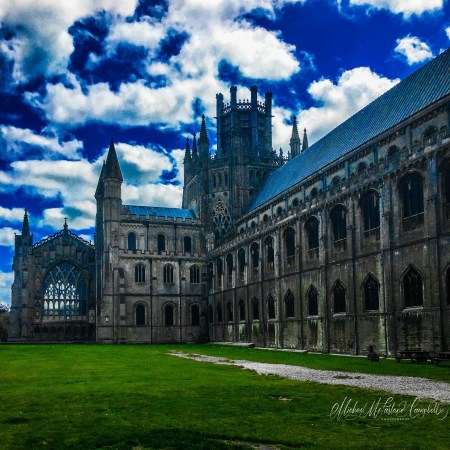 Ely Cathedral rises above a green close beneath towering clouds, its ancient stonework framed by light, shadow, and a vivid blue sky.