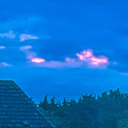 A dramatic blue sky streaked with pink light rises above a tiled roof and dark trees at dusk.