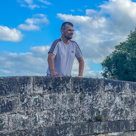 A man in a white T-shirt leans on the stone parapet of a canal bridge, looking into the distance beneath a wide blue sky filled with white clouds.