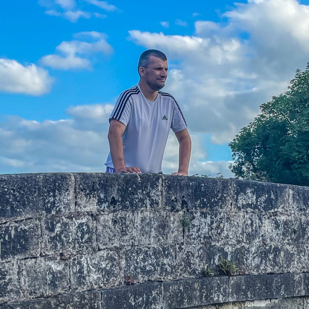 A man in a white T-shirt leans on the stone parapet of a canal bridge, looking into the distance beneath a wide blue sky filled with white clouds.