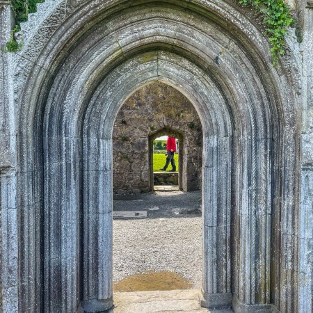 Carved medieval stone arch at Clonmacnoise framing a distant doorway, where a visitor in a red jacket walks across the opening beyond.