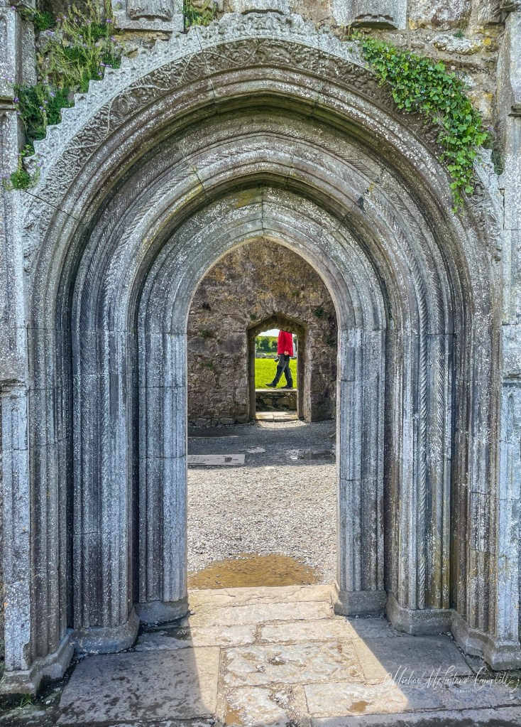 Carved medieval stone arch at Clonmacnoise framing a distant doorway, where a visitor in a red jacket walks across the opening beyond.