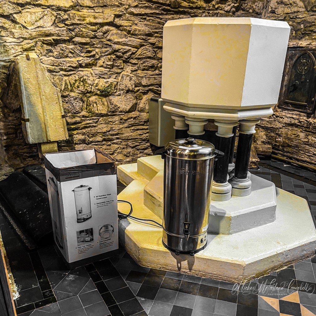 A medieval stone baptismal font stands in a church beside a modern hot-water urn prepared for refreshments after worship.