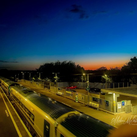 A train stands at Monasterevin railway station at dusk, viewed from an overhead footbridge. Platform lights glow against the deep blue evening sky, while a band of orange sunset lingers on the horizon. A crescent moon hangs above the station as the last light of day fades into night.