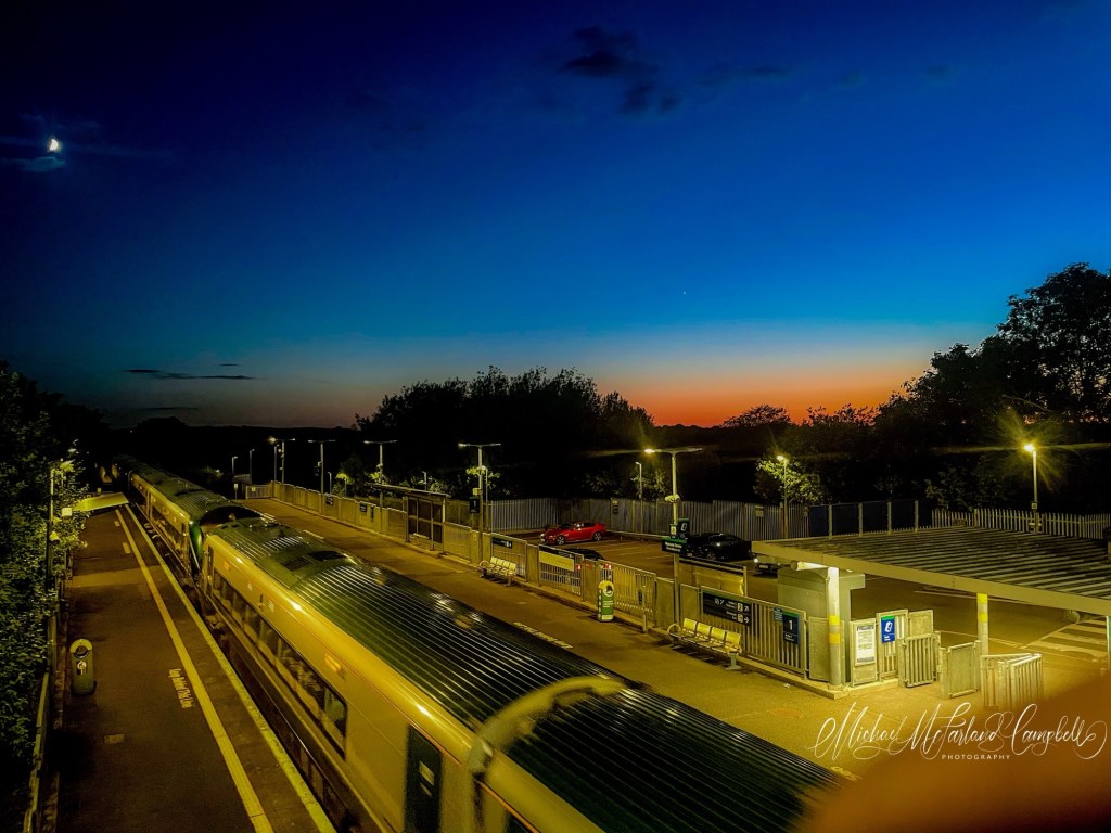 A train stands at Monasterevin railway station at dusk, viewed from an overhead footbridge. Platform lights glow against the deep blue evening sky, while a band of orange sunset lingers on the horizon. A crescent moon hangs above the station as the last light of day fades into night.