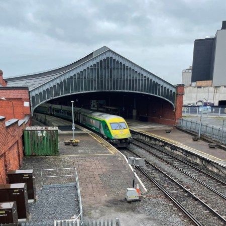 Irish Rail train standing beneath the curved roof of Kent Station in Cork on a cloudy day, with tracks leading into the station.