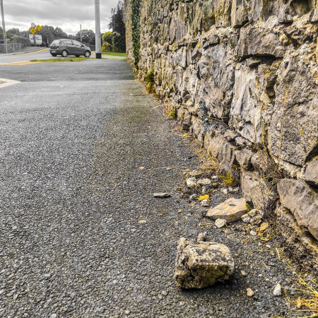 A weathered stone wall in Monasterevin with several loose stones lying on the pavement beside it, photographed from ground level looking along the wall.