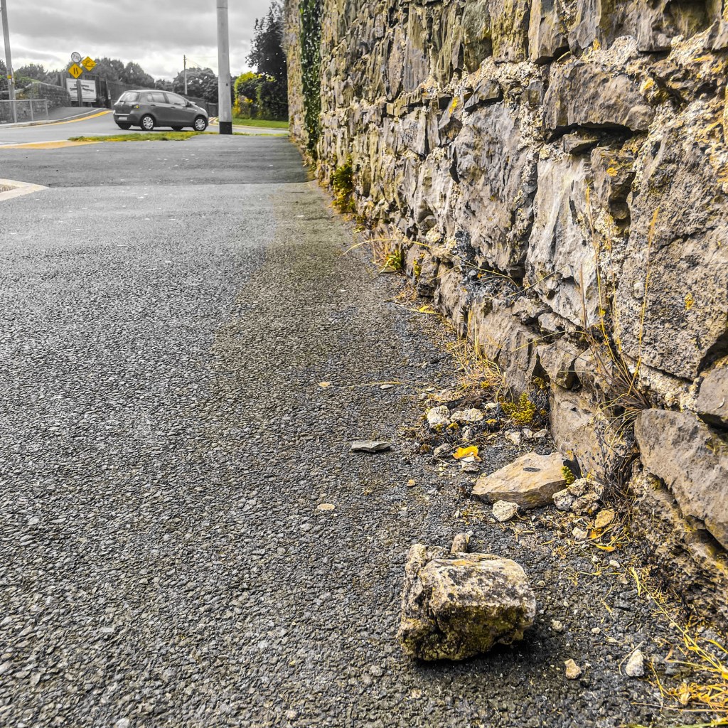 A weathered stone wall in Monasterevin with several loose stones lying on the pavement beside it, photographed from ground level looking along the wall.