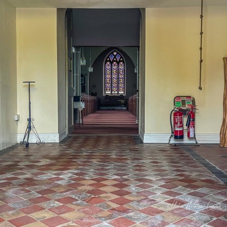 View through a church entrance into the nave, with a patterned tiled floor, stained-glass window and altar ahead, a candle stand on the left, fire extinguishers on the right, and a bell rope hanging nearby.