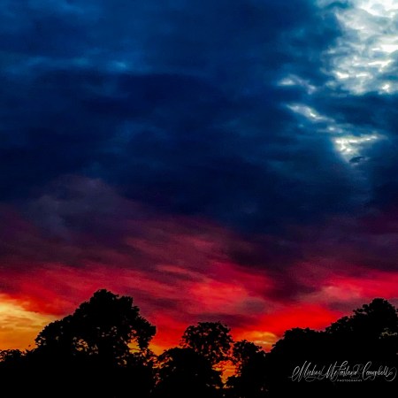 Deep crimson and orange sunset beneath dark blue storm clouds, with silhouetted trees along the horizon in Monasterevin.