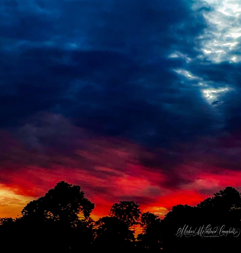 Deep crimson and orange sunset beneath dark blue storm clouds, with silhouetted trees along the horizon in Monasterevin.