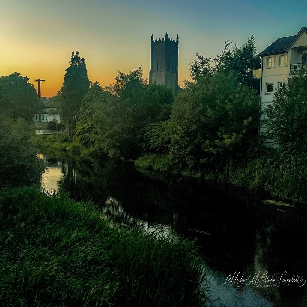 Evening view across the River Barrow in Monasterevin with the church tower silhouetted against a fading golden sky and reflected in the calm water below.