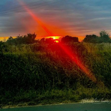 Vivid orange-red sunset over summer fields and hedgerows with a dramatic shaft of sunlight stretching across the sky.