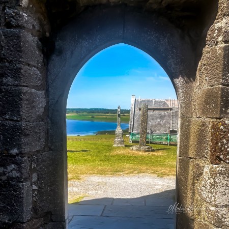 View through the dark stone Whispering Arch at Clonmacnoise Cathedral towards Temple Connor, high crosses, green fields, and the River Shannon.