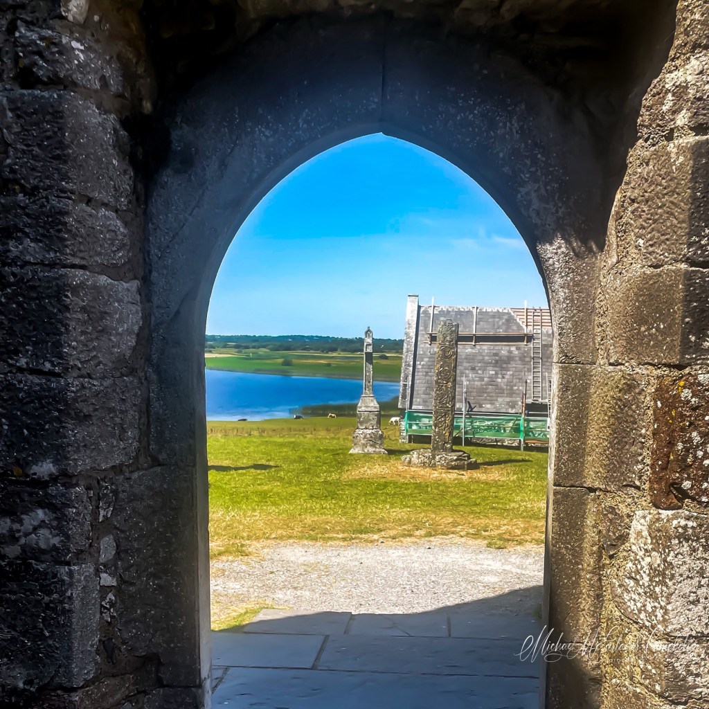 View through the dark stone Whispering Arch at Clonmacnoise Cathedral towards Temple Connor, high crosses, green fields, and the River Shannon.