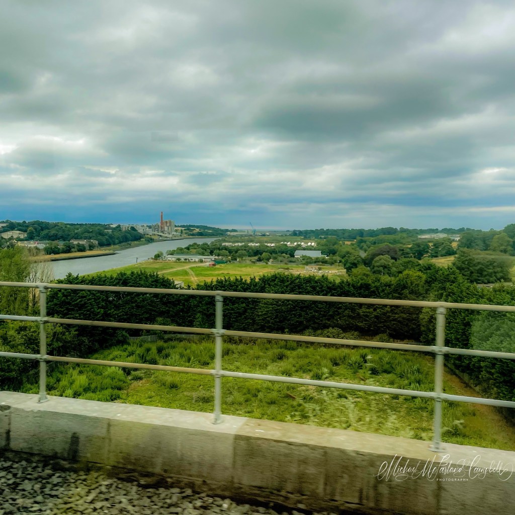 View from a moving train crossing the Drogheda viaduct, looking across the River Boyne beneath a dramatic overcast sky, with green fields, trees, and the town in the distance framed by the railway parapet.