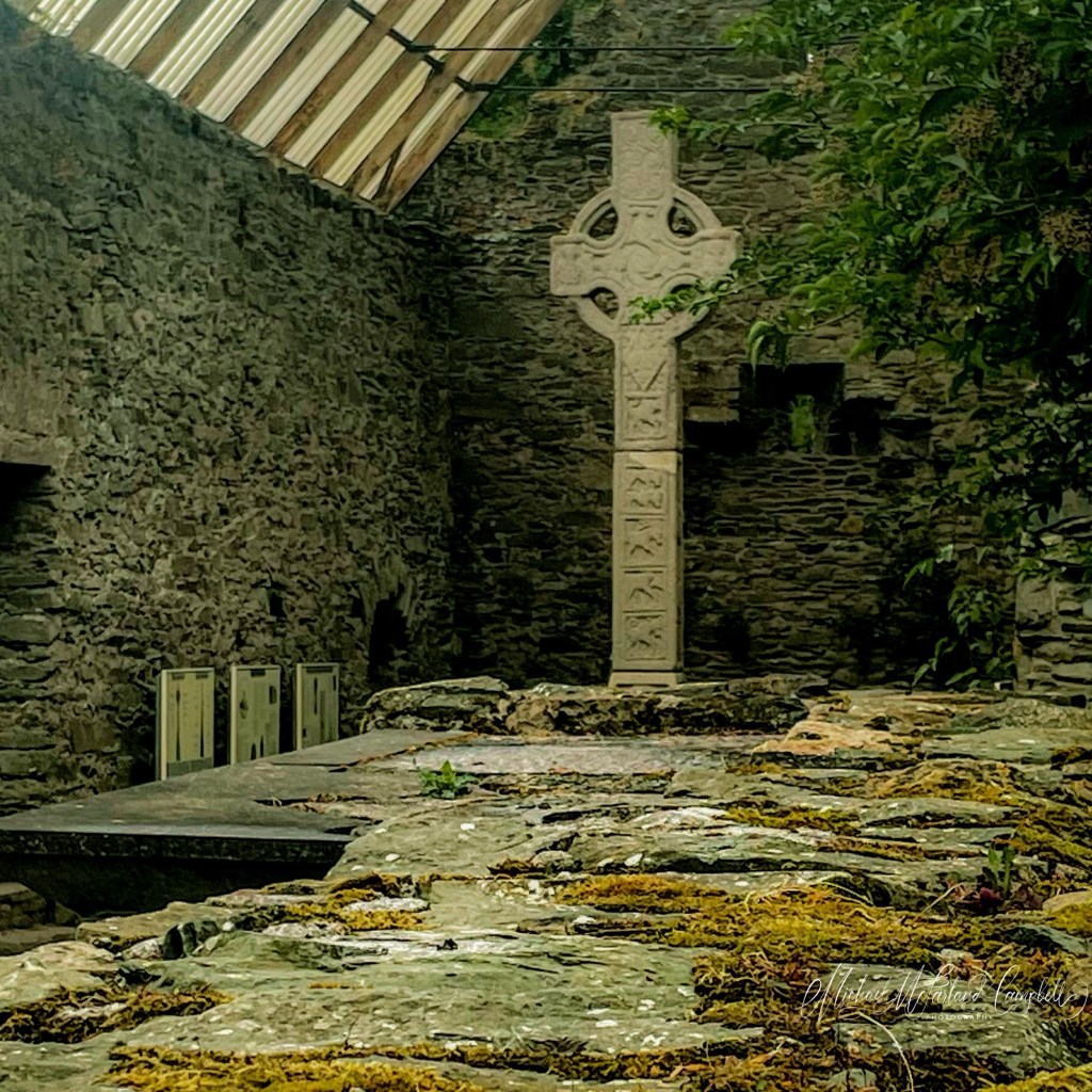 Moone High Cross standing within the ruins at Moone, County Kildare, seen across ancient stone slabs mottled with moss. The pale carved cross rises against the dark stone wall, with leafy branches entering from the right and part of the protective roof visible above.