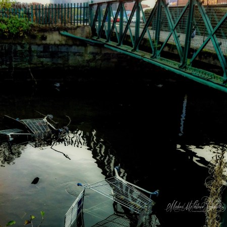 Several shopping trolleys partly submerged in dark water beneath a green metal bridge, surrounded by rippling reflections and stone walls.