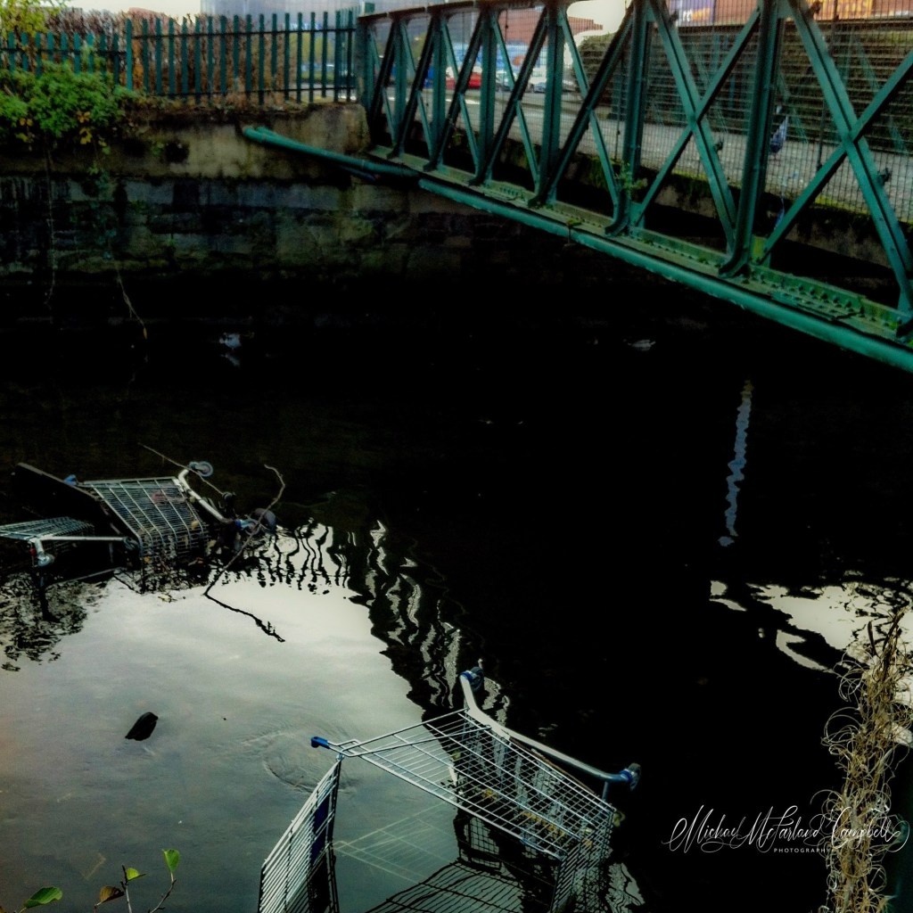 Several shopping trolleys partly submerged in dark water beneath a green metal bridge, surrounded by rippling reflections and stone walls.