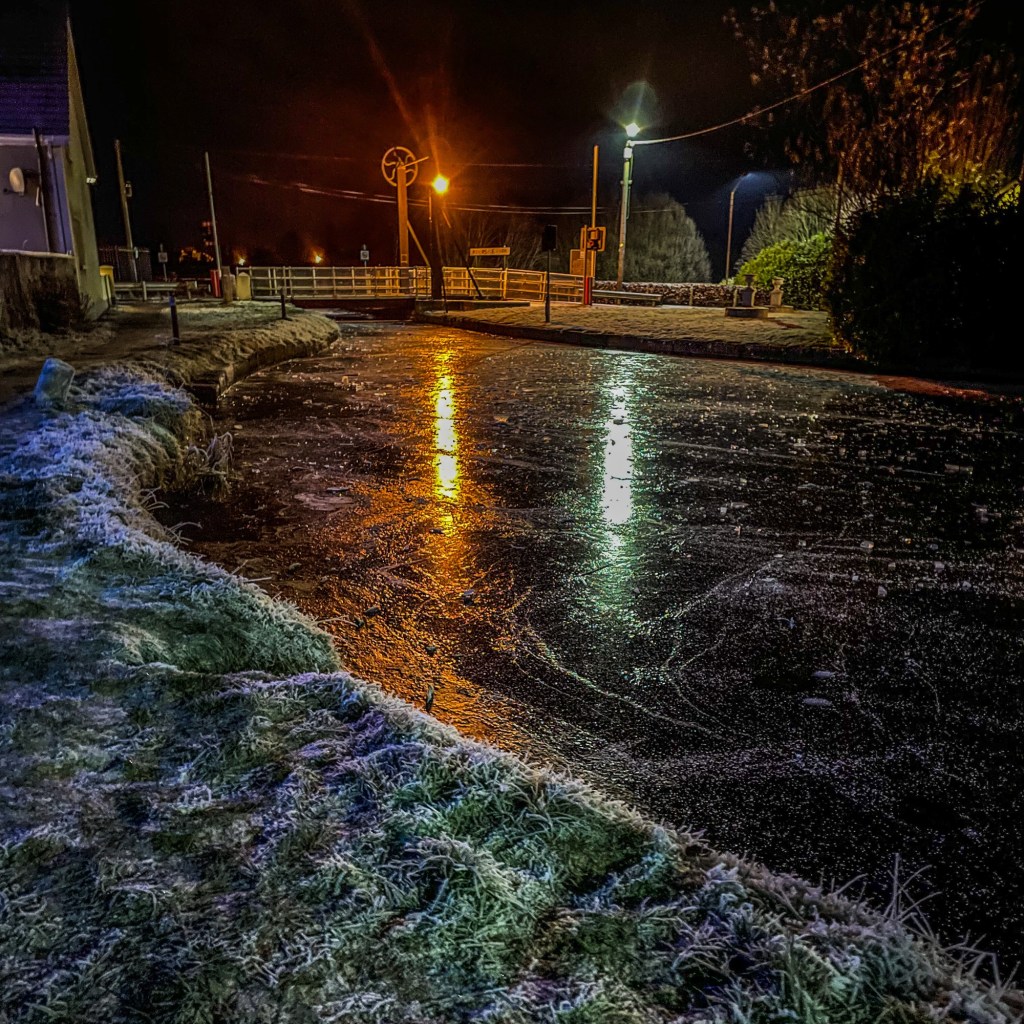 Frost-covered grass beside a dark canal, with amber and white reflections leading towards a drawbridge beneath streetlights.