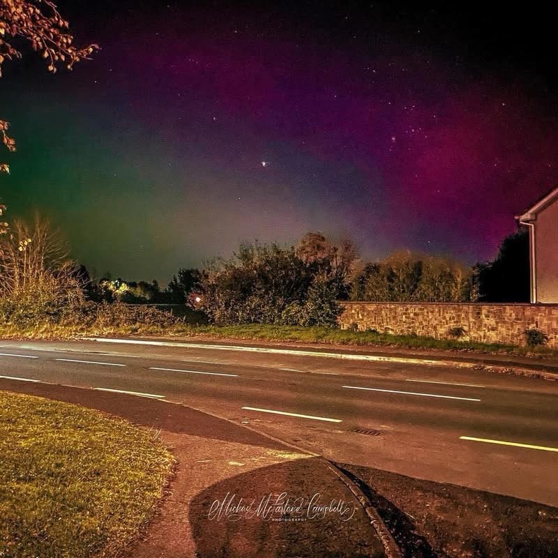 Green and purple aurora across a starry night sky above trees, a stone wall and a house beside a road in Monasterevin.